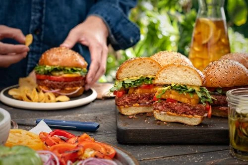 Plant-based burger still life on a wood table, featuring an adult holding a chip, with leafy background.