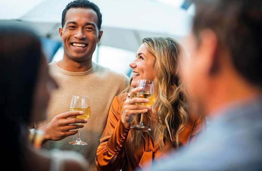 Smiling man and woman holding wine glasses at an outdoor social gathering, with blurred people in the foreground and background.