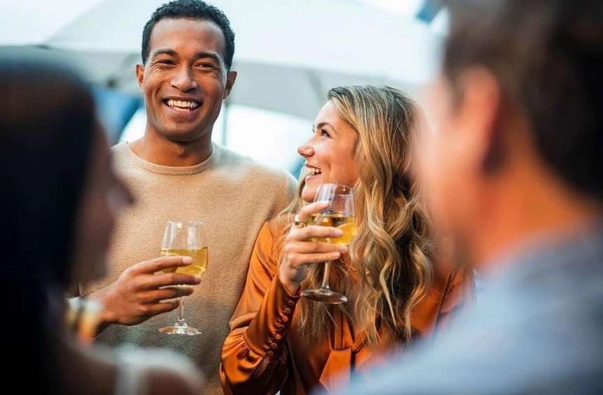 Smiling man and woman holding wine glasses at an outdoor social gathering, with blurred people in the foreground and background.
