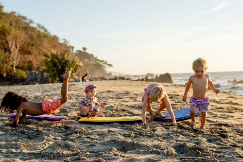 Four young children play on a sandy beach with surfboards at sunset. One child has legs in the air.