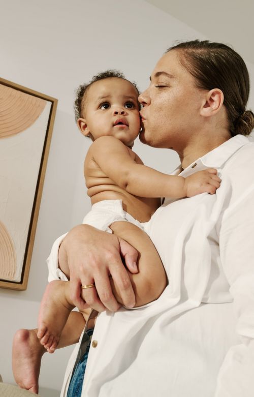 Mother kissing her baby on the forehead indoors. The baby is wrapped in a diaper, held in her mother's arms. Simple wall art in the background.