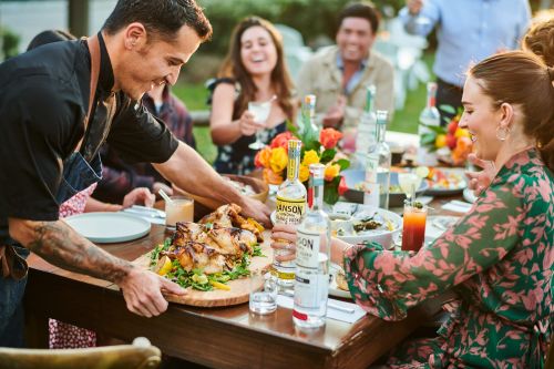A catered outdoor dining experience with a server presenting roasted chicken to a cheerful group of adults at a long table.