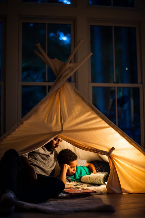 Father and child bonding, reading inside a cozy tent illuminated with warm light, creating a heartwarming indoor family scene.