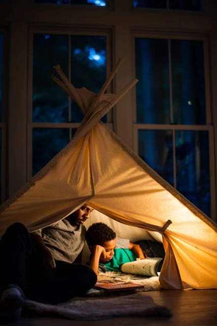 Father and child bonding, reading inside a cozy tent illuminated with warm light, creating a heartwarming indoor family scene.