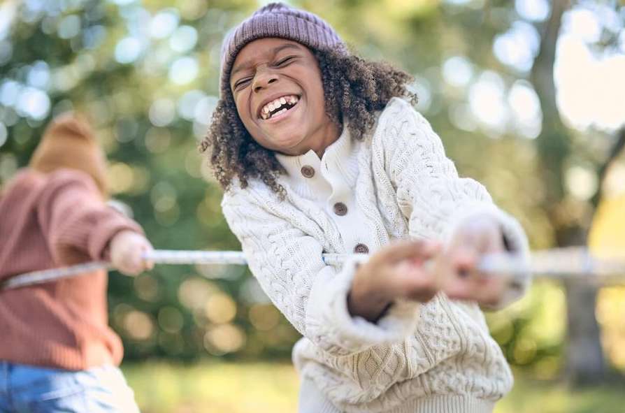 A laughing child with curly hair wearing a knitted hat and sweater participates in tug-of-war outdoors on a bright, sunny day.
