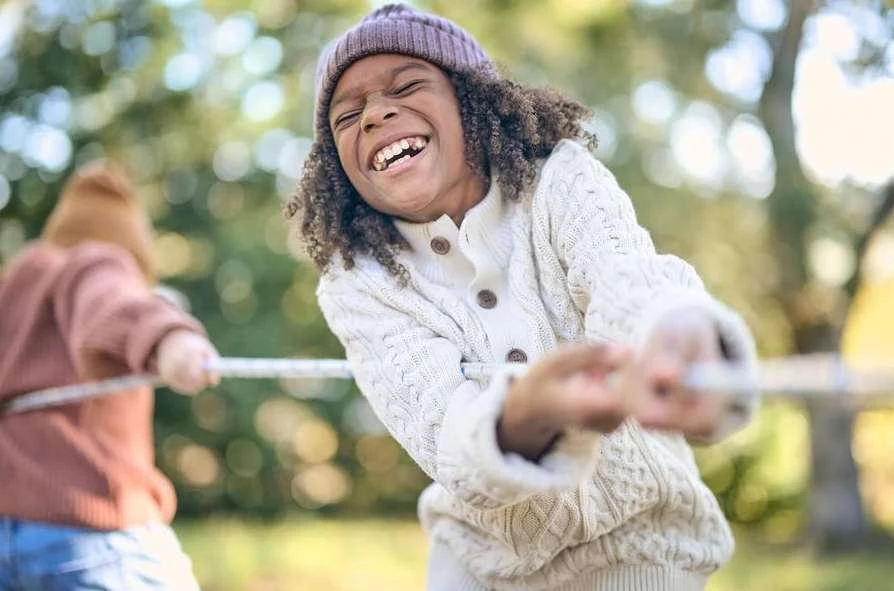 A laughing child with curly hair wearing a knitted hat and sweater participates in tug-of-war outdoors on a bright, sunny day.
