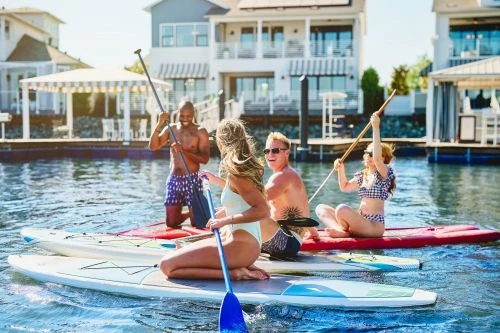 Group of four people paddleboarding in a waterway near waterfront homes on a sunny day; active outdoor lifestyle in a residential setting.