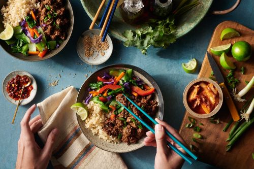 Overhead shot showcases hands using chopsticks to eat Korean beef bowls with rice, vegetables, limes, kimchi, and spices on a blue table.