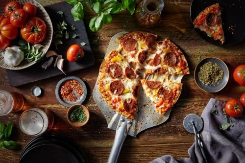 Overhead shot of a pepperoni pizza on a metal peel, surrounded by tomatoes, garlic, herbs, and spices on a wooden surface.