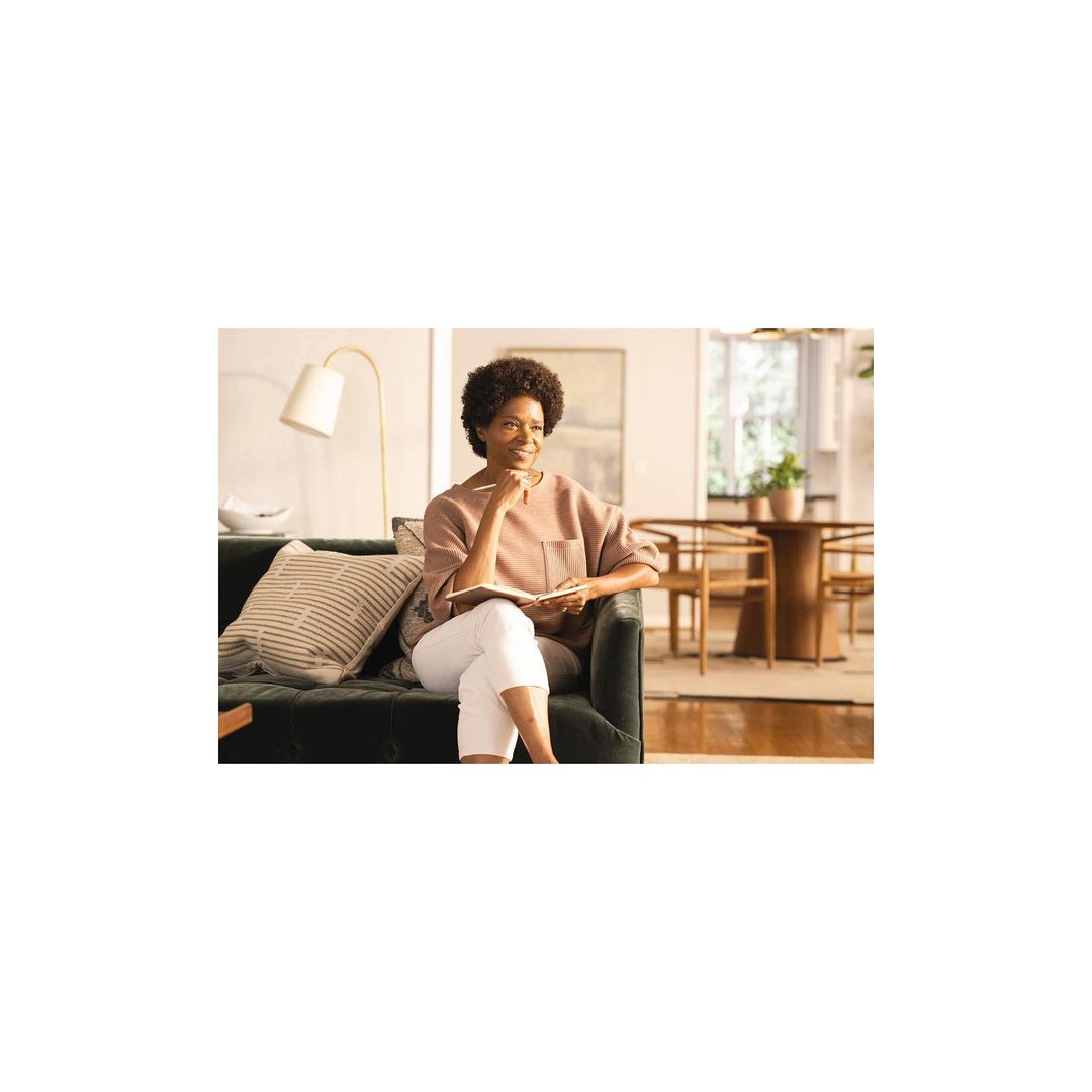 A woman sits on a green velvet sofa, smiling and holding a notebook. Natural light fills the living room with a modern interior.
