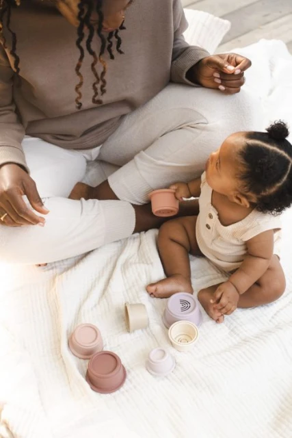 A toddler and an adult sit on a white blanket with a collection of pastel-colored silicone stacking cups during playtime.