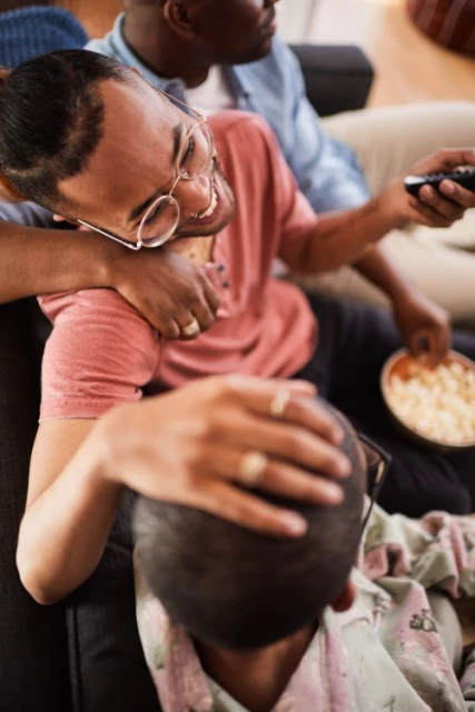 Three African-American men relax on a couch while watching TV, one holds the remote, another has a snack bowl in a bright living room.