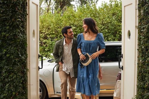 Attractive couple poses in front of a white car and lush greenery. The woman is wearing a blue dress and the man is wearing a green jacket.