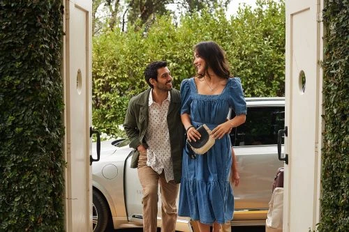 Attractive couple poses in front of a white car and lush greenery. The woman is wearing a blue dress and the man is wearing a green jacket.