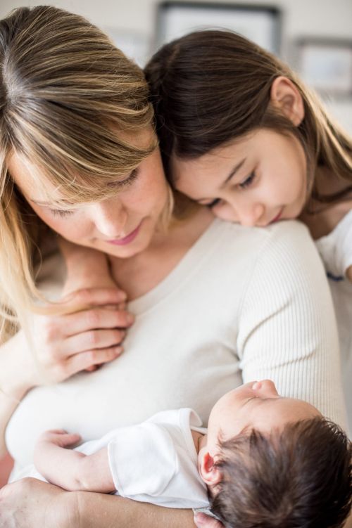 Close-up of a mother holding a baby with a girl resting her head on the mother's shoulder, indoor setting, soft lighting, and neutral colors.