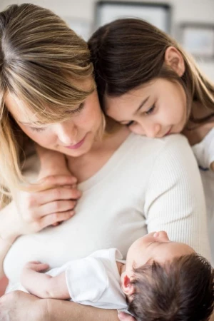 Close-up of a mother holding a baby with a girl resting her head on the mother's shoulder, indoor setting, soft lighting, and neutral colors.