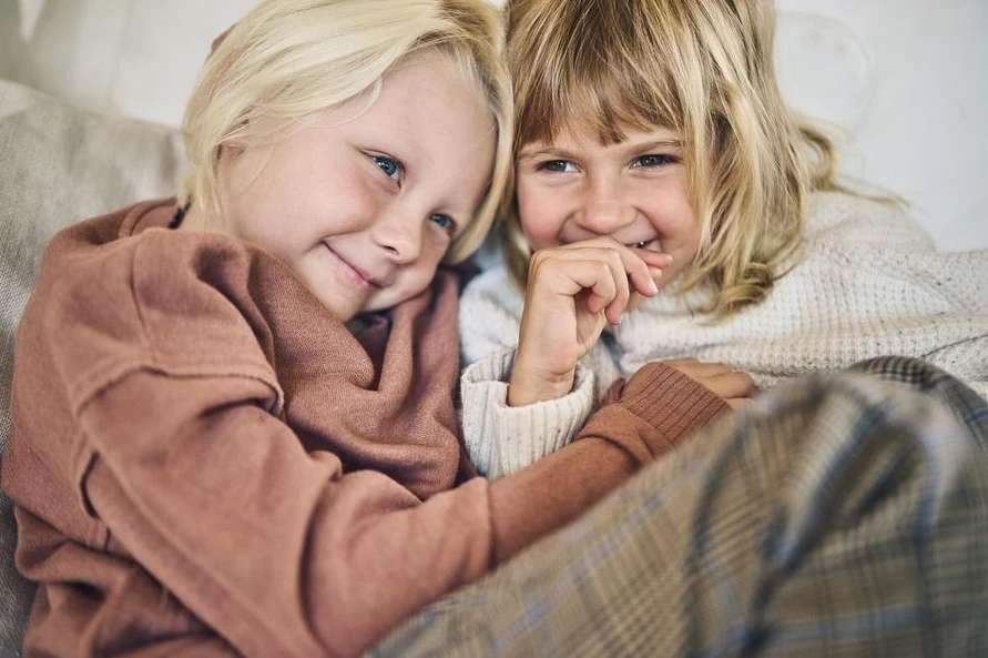Two happy young girls with blonde hair are indoors, sitting closely together wearing casual sweaters and plaid pants, smiling and laughing.