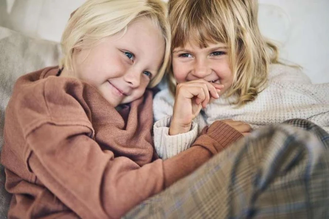 Two happy young girls with blonde hair are indoors, sitting closely together wearing casual sweaters and plaid pants, smiling and laughing.