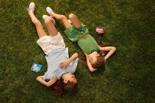 Two young girls lie on green grass in an overhead shot, wearing casual summer clothes with natural light illuminating the scene.