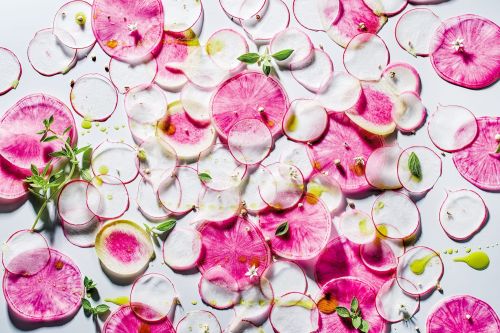 Overhead shot of thinly sliced radishes arranged on a light surface with small green leaves and glistening olive oil droplets.