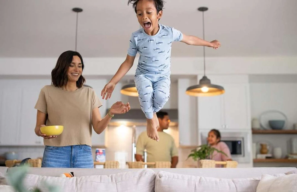 A young boy joyfully leaps over a sofa in a brightly lit home interior, while an adult female and child look on in the background.