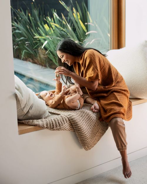 A mother in orange dress smiles while playing with her baby on a windowsill with pillows and blanket, with a pool visible outside.