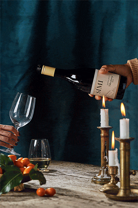 Pouring white wine into a glass next to lit candles, accompanied by citrus fruits on a wooden table against a dark teal background.