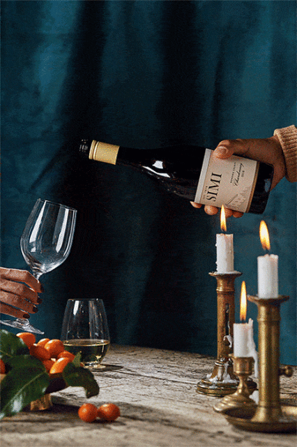 Pouring white wine into a glass next to lit candles, accompanied by citrus fruits on a wooden table against a dark teal background.