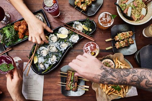 Overhead shot of a wooden table laden with plates of appetizers, skewers, oysters, french fries, and cocktails, showcasing a diverse food spread.