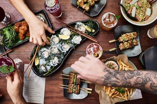Overhead shot of a wooden table laden with plates of appetizers, skewers, oysters, french fries, and cocktails, showcasing a diverse food spread.