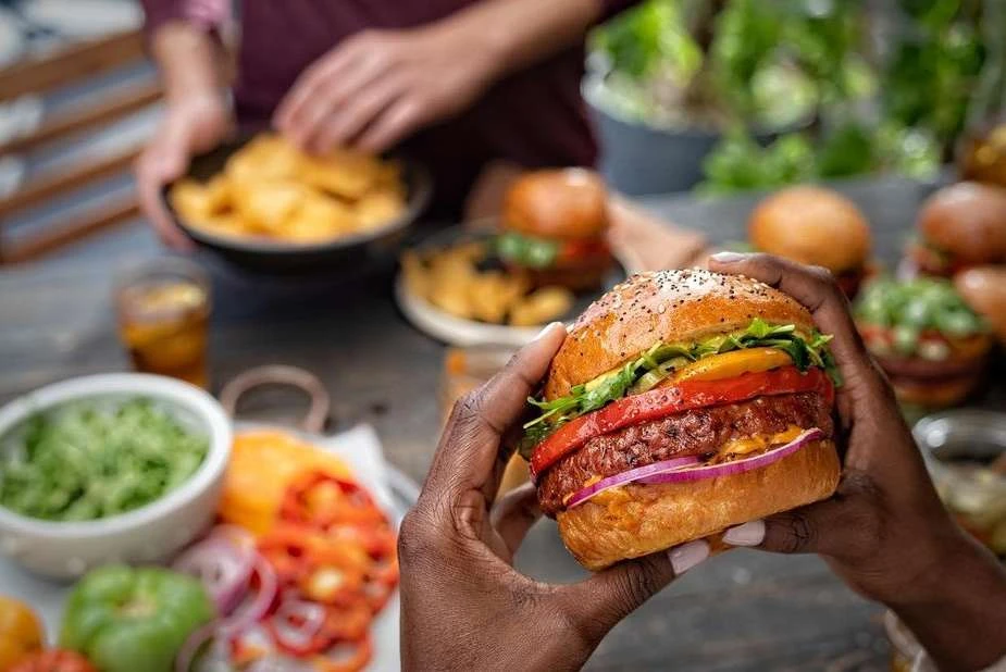 Close-up of a plant-based burger held by a person with dark skin, adorned with fresh toppings and a seeded bun; additional food items on a table.