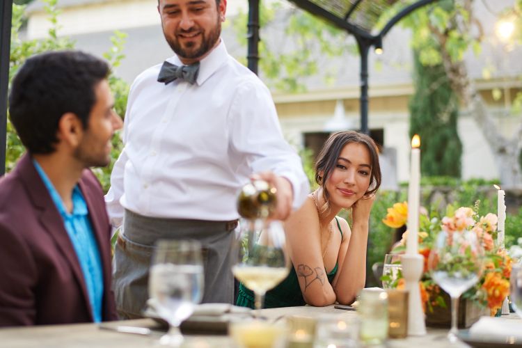 People enjoying a wine pouring at an outdoor event with floral centerpieces and candles on a wooden table in the daytime.
