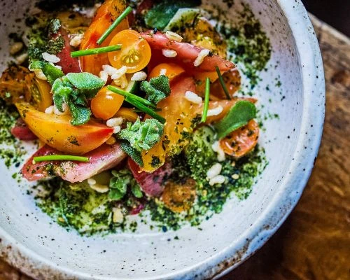 Overhead shot of a colorful salad of sliced heirloom tomatoes, herbs, and grains arranged in a white ceramic bowl on a wood surface.