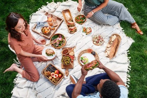 Overhead shot of two adults enjoying a picnic in a grassy outdoor space with various food items on a white striped blanket.