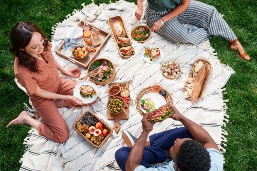 Overhead view of three people having a picnic on a white blanket in the grass, surrounded by sandwiches, salads, fruit, and drinks.