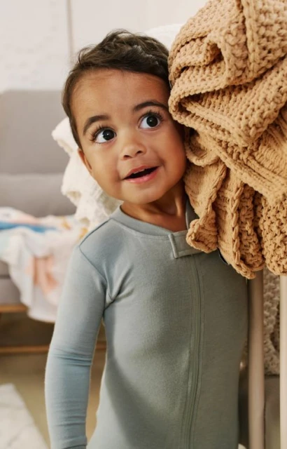 A toddler in pajamas leans against a pile of soft, neutral-toned blankets inside a home, looking off-camera with a joyful facial expression.