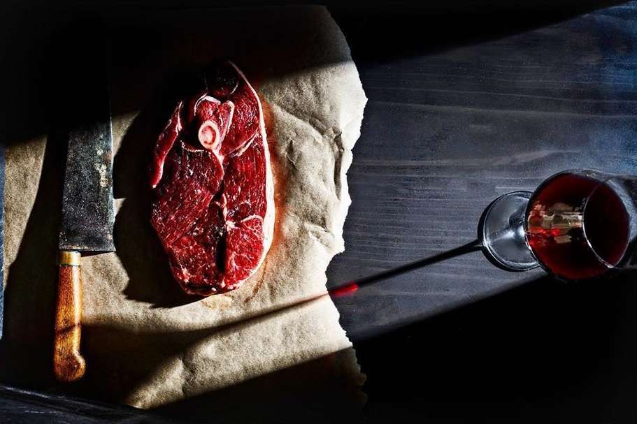 A high-angle shot showcases a raw meat cut, knife, and wine glass arranged on crumpled paper against a dark backdrop under studio lighting.