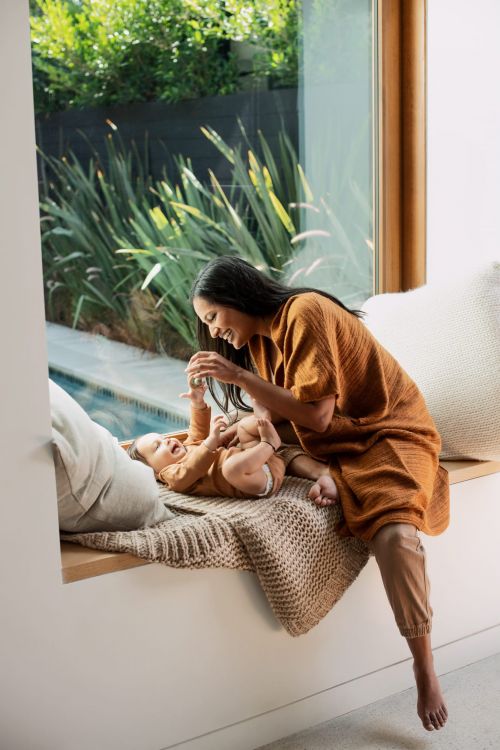 An adult woman in a rust-colored dress interacts with a baby on a woven blanket near a window and a sunlit outdoor patio with greenery.