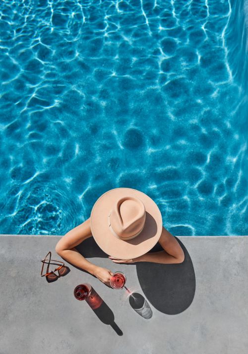 Overhead shot of a woman relaxing by a pool with drinks, sunglasses, and a wide-brimmed hat, enjoying a sunny day.