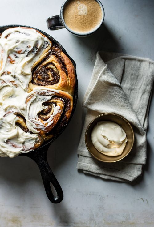 Overhead shot of cinnamon rolls with frosting in a cast iron skillet, with a coffee cup and towel creating an inviting, warm scene.