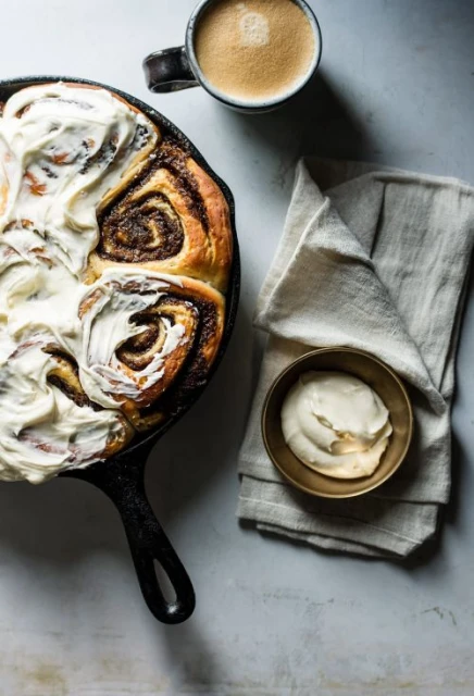 Overhead shot of cinnamon rolls with frosting in a cast iron skillet, with a coffee cup and towel creating an inviting, warm scene.