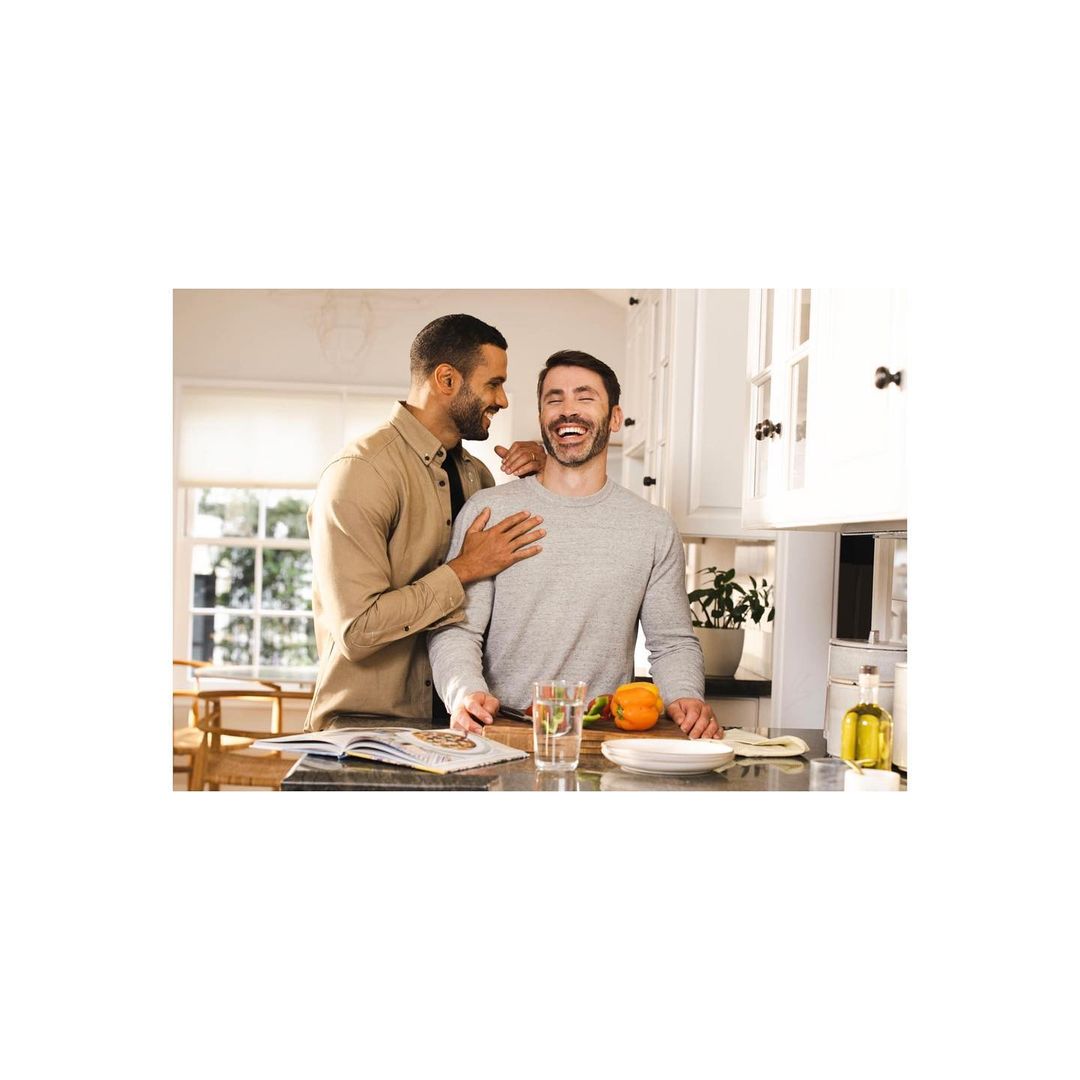 Two men in a bright kitchen, one with his hand on the other's shoulder. Open cookbook, fruit, and glasses on the counter. Light and airy setting.