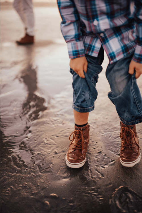 Child in brown boots and denim jeans plays in the wet sand at the beach, while another person stands in the background.