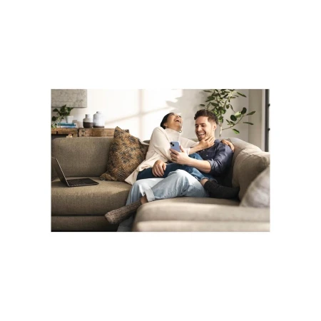 A happy couple relaxes on a couch looking at a smartphone, with a laptop nearby, indoor plants, and natural light.