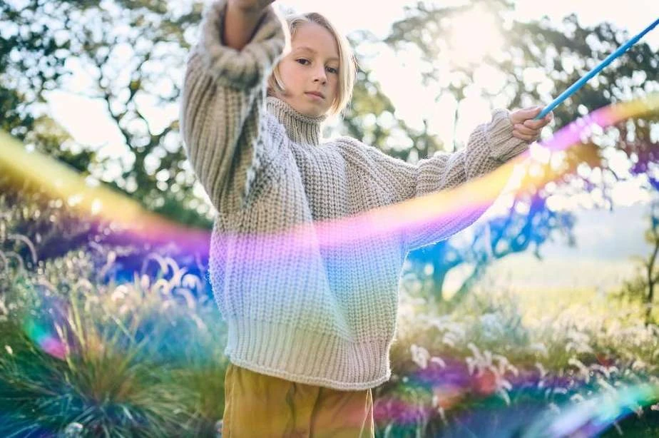 Young child in chunky knit sweater holds blue string in a sunny, outdoor environment with lush greenery, blurred background, rainbow bokeh.