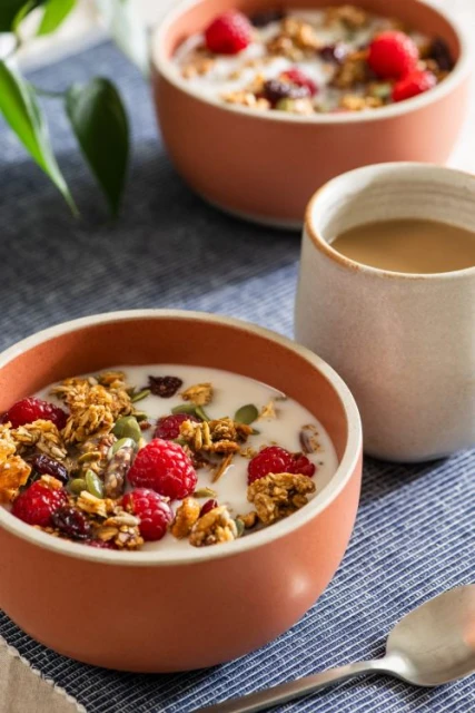 Close-up of two bowls filled with granola, raspberries, and milk, next to a cup on a blue patterned placemat with natural lighting.