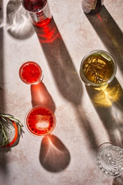 Overhead view: cocktails with ice, rosemary garnish, and bottles on a textured surface with shadows, emphasizing a vibrant and refreshing beverage concept.