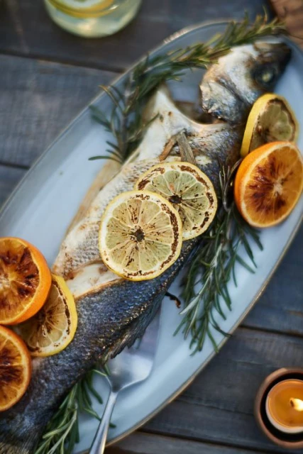 Overhead shot of grilled sea bass plated with lemon and orange slices, garnished with rosemary sprigs on a rustic wooden table.
