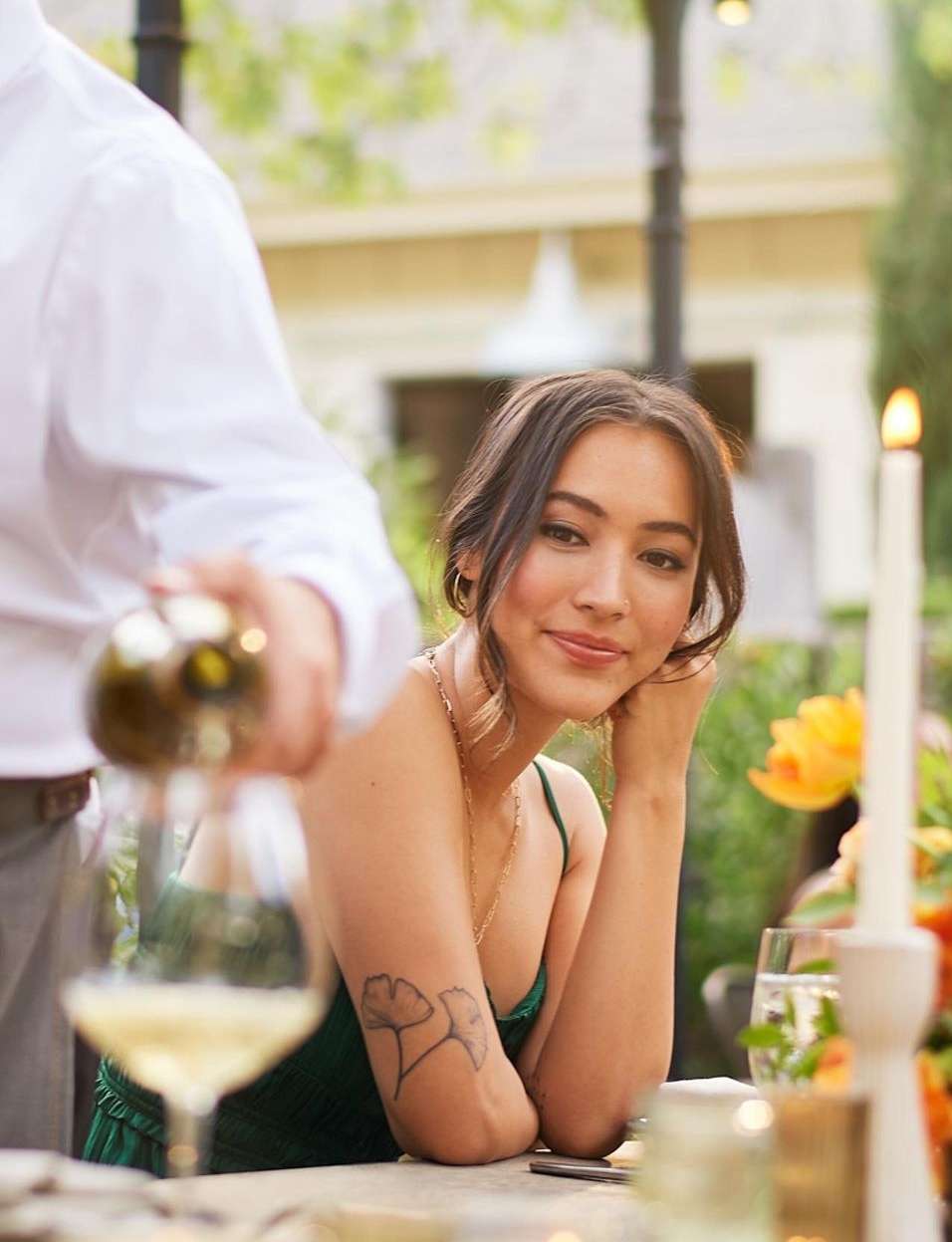 Young woman in green dress seated at a table as someone pours white wine, creating a relaxed outdoor dining scene with soft lighting.