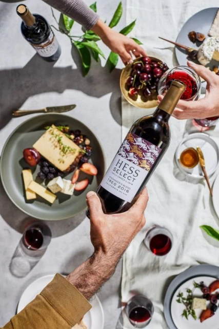 Overhead shot of a wine bottle being held over a table with cheese, fruit, and wine glasses, creating an inviting food and beverage scene.